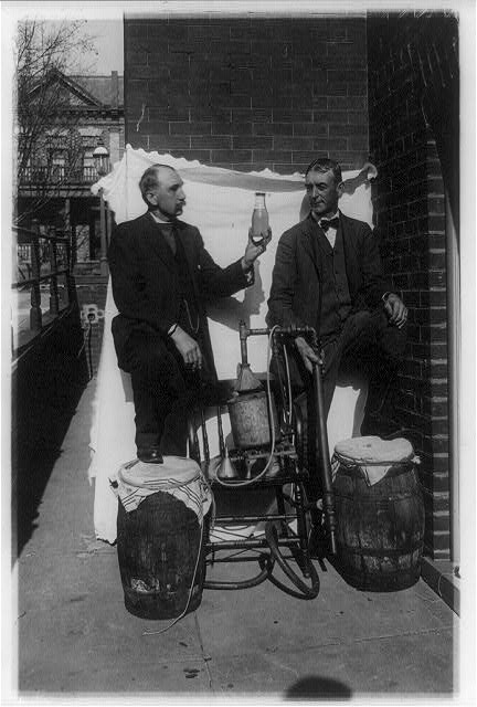 Two men standing outdoors beside a small still; one holds a bottle of liquor up toward the camera.