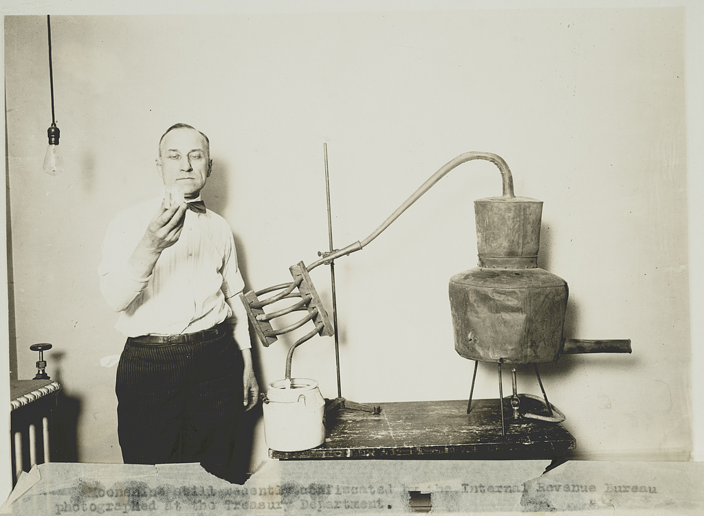 A man in shirtsleeves stands beside a copper still on a table, holding a glass up to look at its contents.