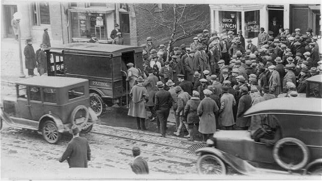 A cropped view of the same raid: a crowd around a police patrol wagon marked Patrol beside parked automobiles on a city street with streetcar tracks