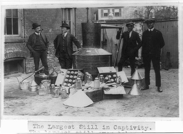 Four men in hats and coats stand around a large still, with jars, funnels and bottles of liquor laid out on the ground in a yard.