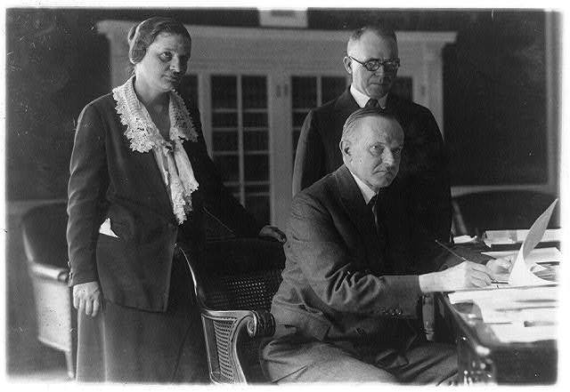 A seated president holding a pen at a desk with a woman and a man standing behind him.