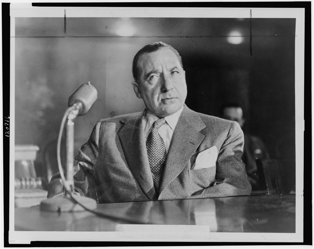 Half-length photograph of Frank Costello seated behind a microphone at a Kefauver Committee hearing.