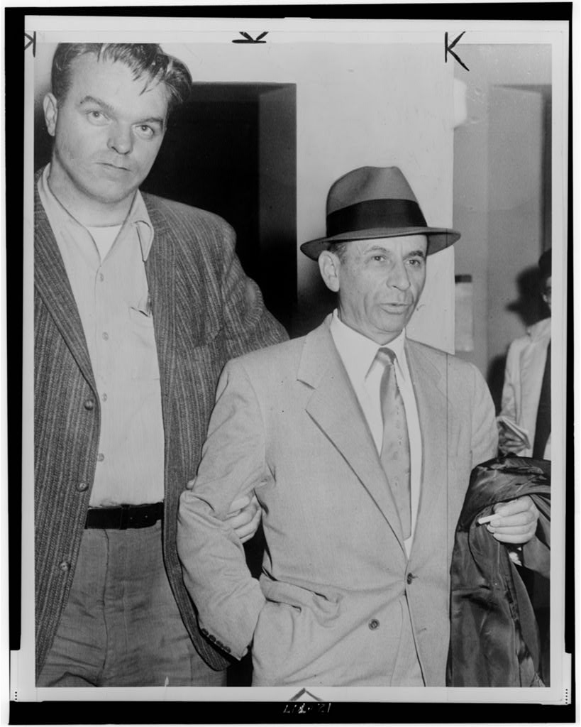 Half-length photograph of Meyer Lansky being led by a detective at a New York City police station.