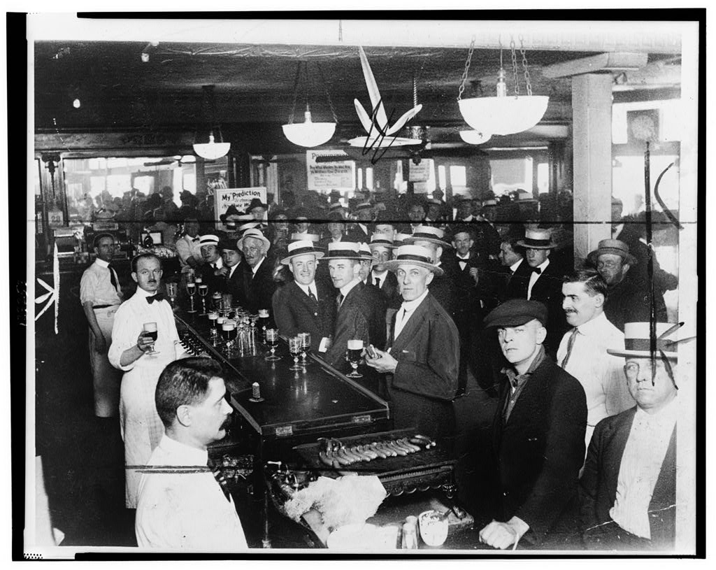 Customers standing shoulder to shoulder along a bar with a bartender behind it, in a New York City saloon interior.