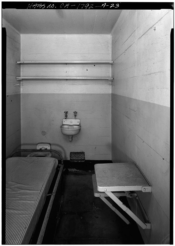 Black-and-white HABS photograph of a small Alcatraz cell with a bunk at the left, a fold-down table at the right, a shelf above and a sink at the back.