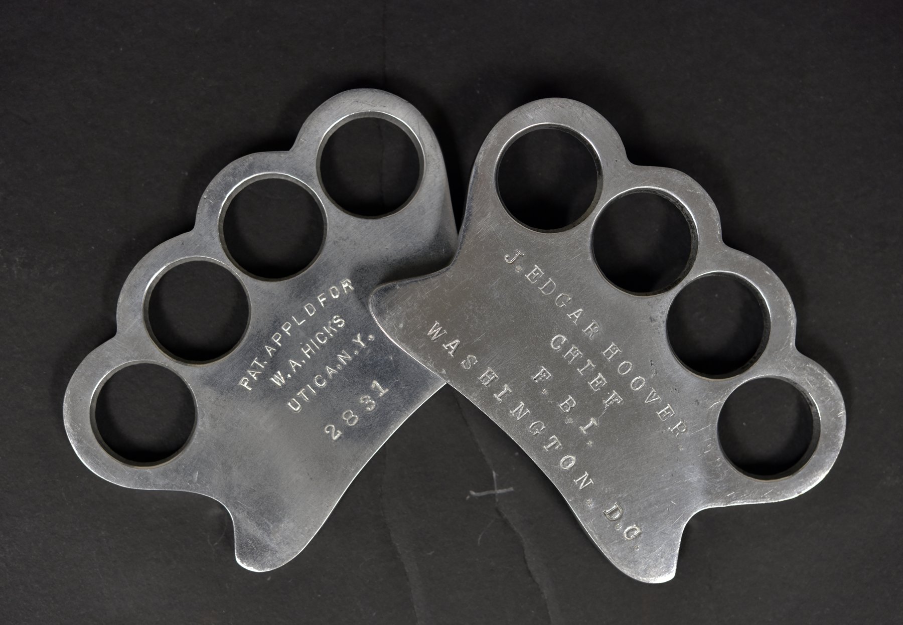 Two metal knuckle-dusters on black cloth; one is stamped Pat. Appl'd For W. A. Hicks Utica N.Y. 2831 and the other is engraved J. Edgar Hoover Chief F.B.I. Washington D.C.