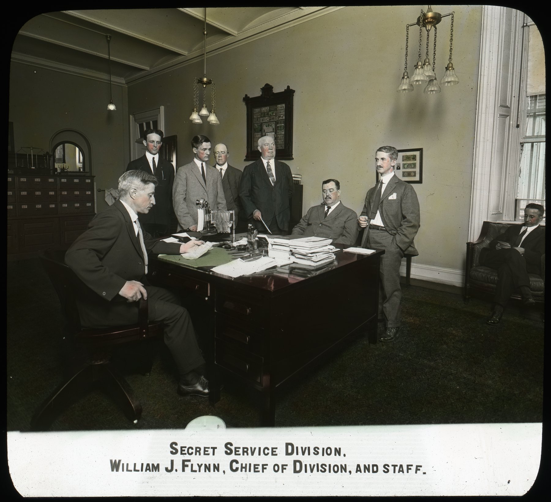 Hand-coloured lantern slide of a laboratory: a long bench with rows of glass flasks and condensers above labelled whiskey bottles, a bald man in a vest standing at the right, and the printed caption Internal Revenue Bureau, Chemical Laboratory, Testing Whiskies.