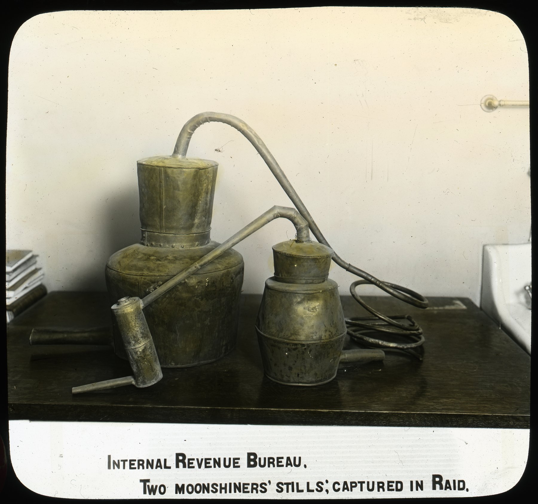 Hand-coloured lantern slide of two small metal pot stills with coiled copper tubing standing on a table, under the printed caption Internal Revenue Bureau. Two Moonshiners' Stills; Captured in Raid.