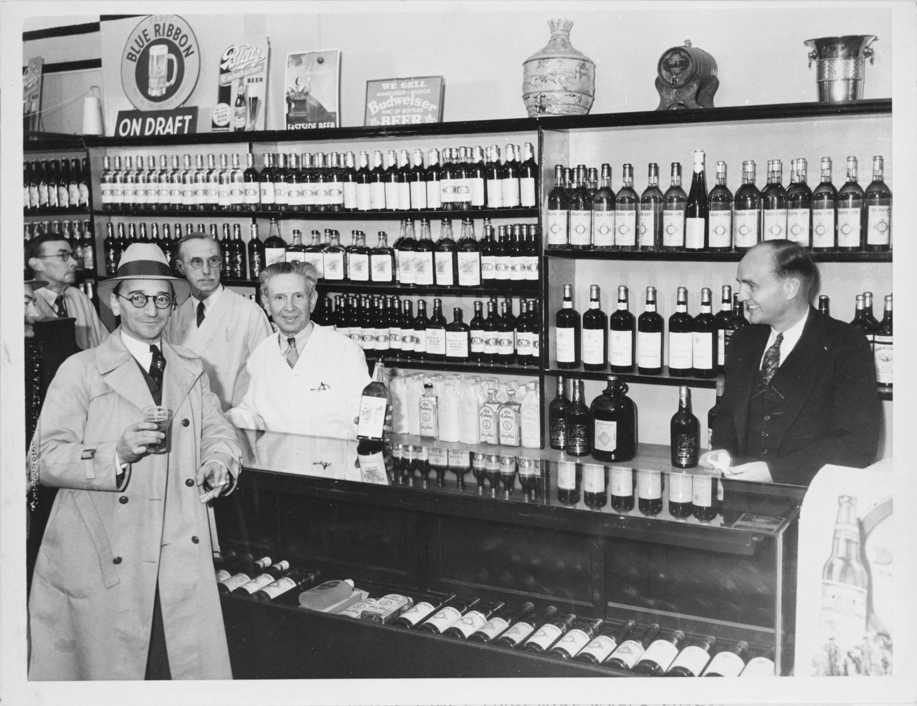 A liquor store counter with shelves of bottles and signs for Blue Ribbon on draft and Budweiser beer; a man in a raincoat and hat holds a small glass at left and four men stand behind and beside the counter.
