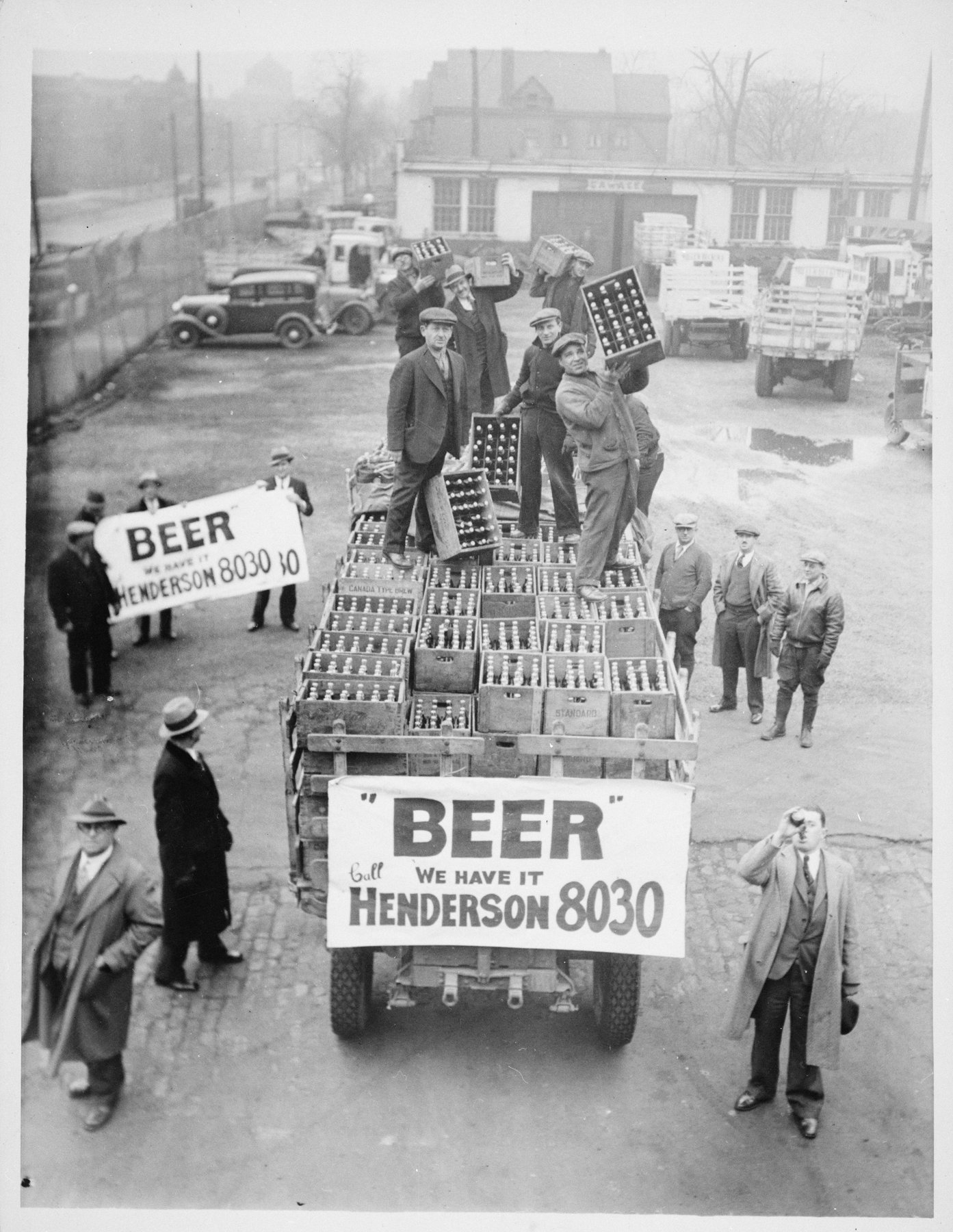 High view of a yard where men stand on a truck stacked with wooden crates of bottles and lift them down; banners on the truck read Beer, We have it, and a man at right drinks from a bottle.