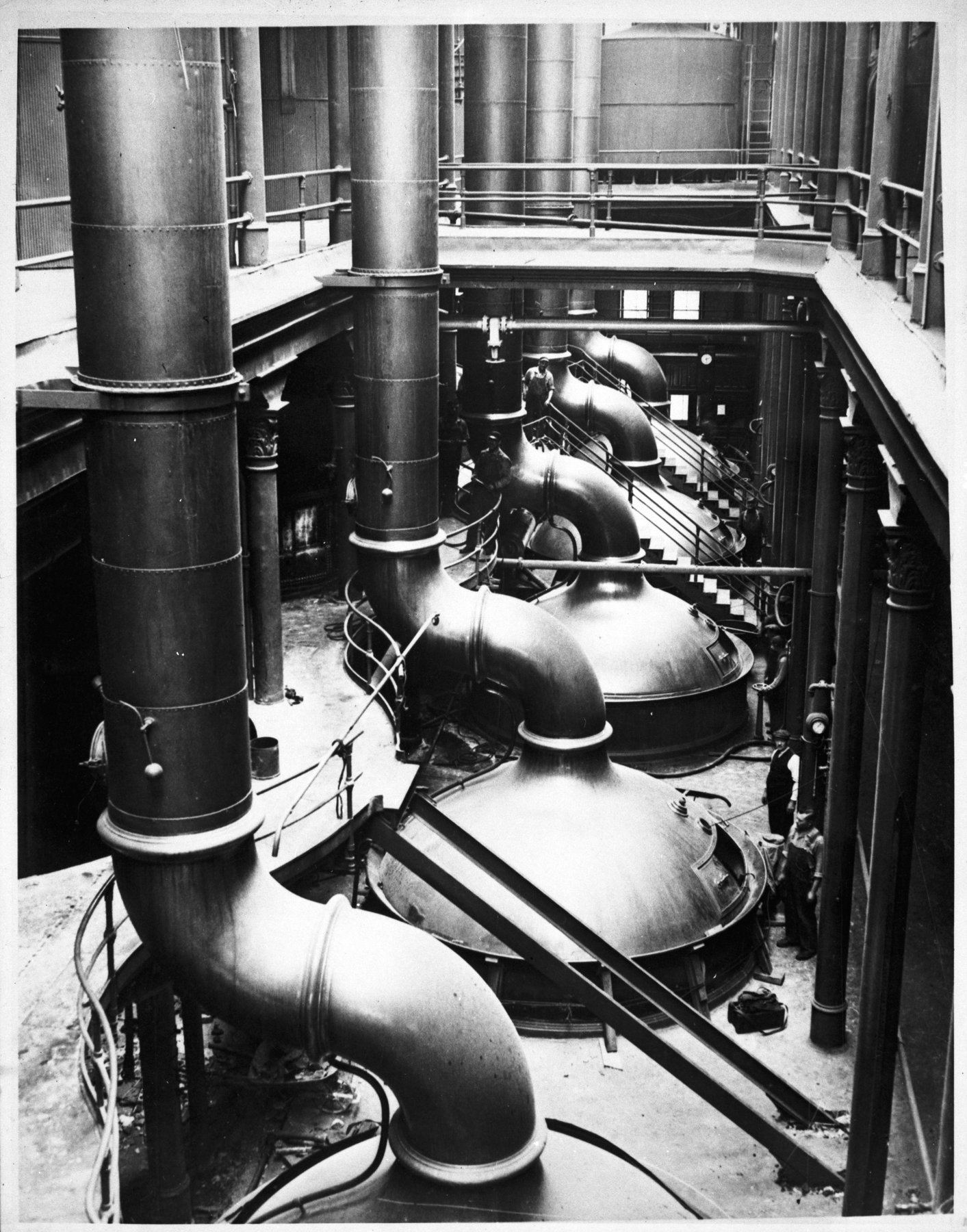 Interior of a brewhouse with a row of large domed kettles, curved vent pipes, stairs and railings, and a few workmen.