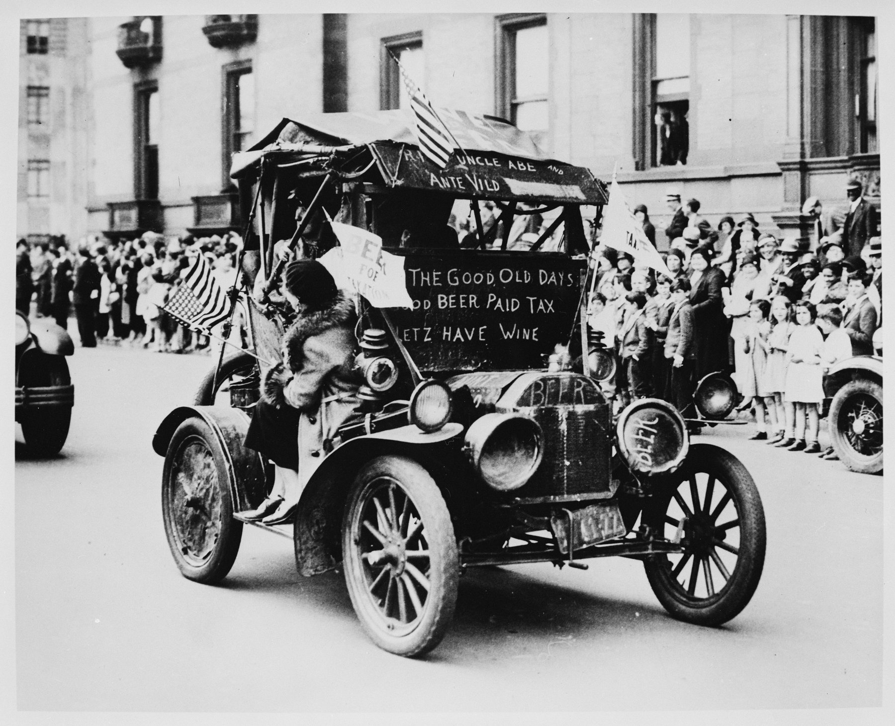 An open early automobile with flags and pennants driving past a crowd on a sidewalk; painted signs on the car refer to the good old days, beer that paid tax, and wine, and Beer is painted on the radiator.