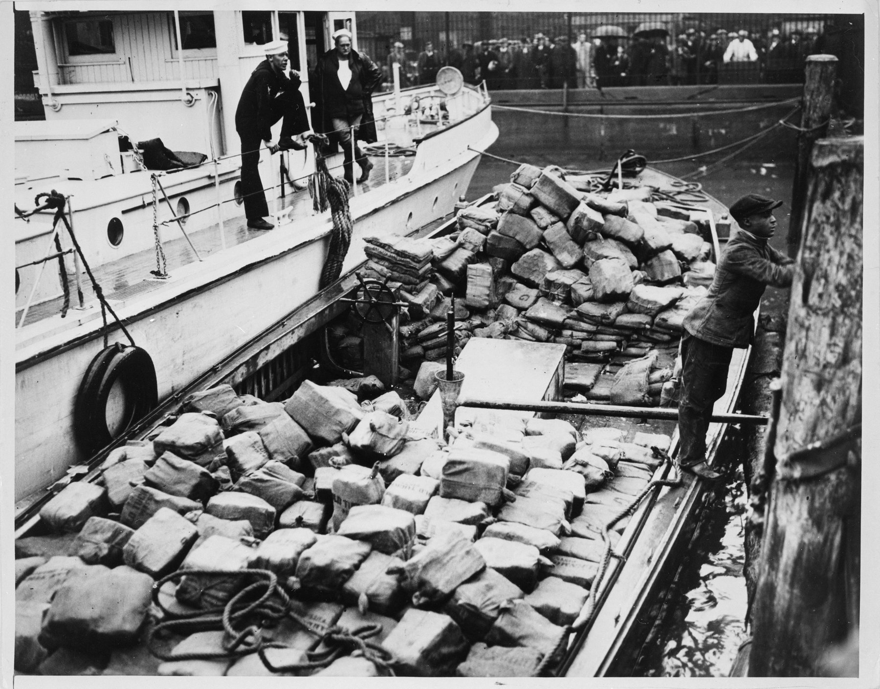 Two boats at a pier: a white vessel with sailors in white caps on deck alongside an open boat heaped with burlap-wrapped bundles, and a crowd behind a fence on the pier.