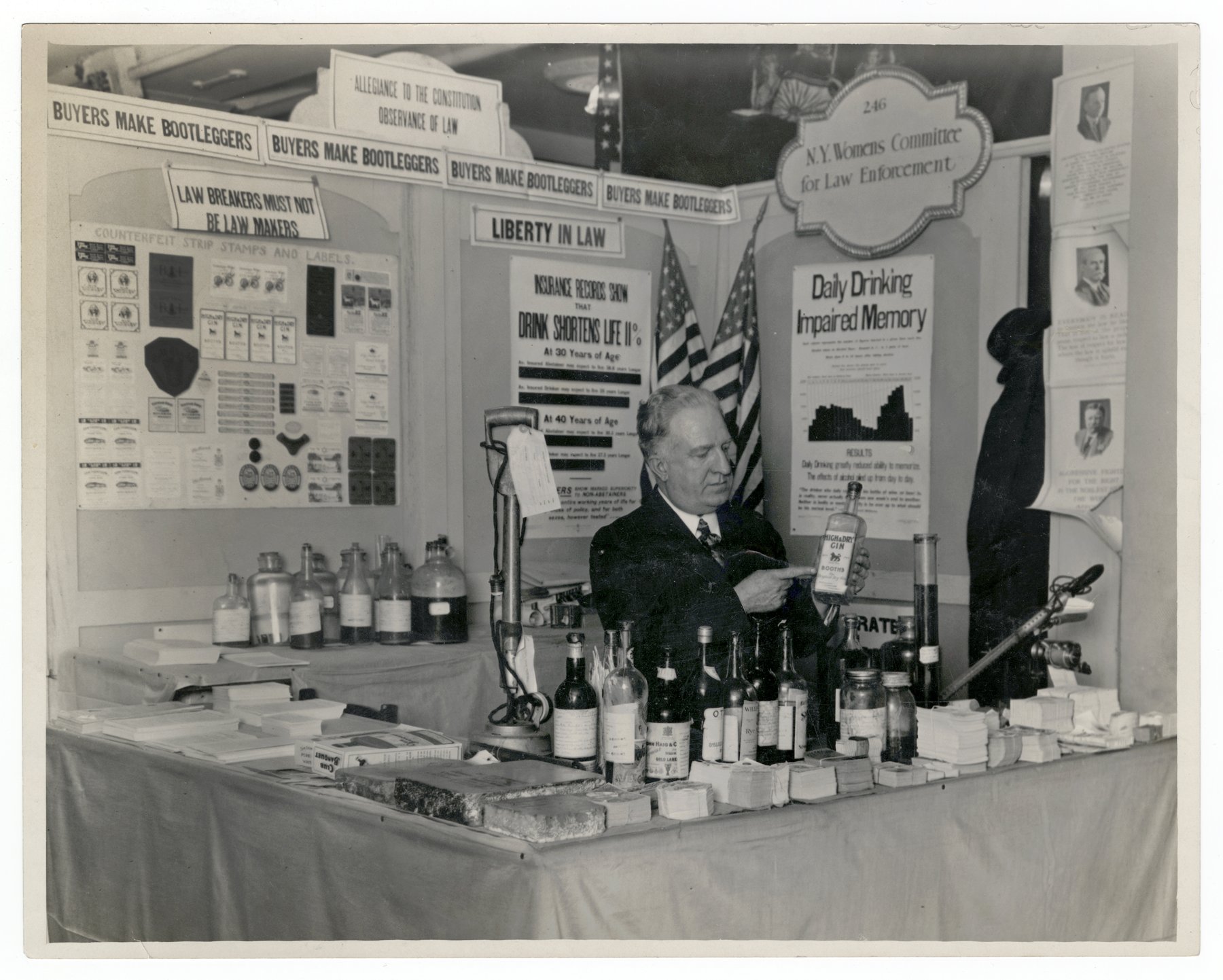 An exhibition booth with signs reading Buyers Make Bootleggers, Allegiance to the Constitution, Law Breakers Must Not Be Law Makers, Liberty in Law and Daily Drinking Impaired Memory, a board of counterfeit strip stamps and labels, flags, and a man behind a table of bottles holding a bottle of gin.