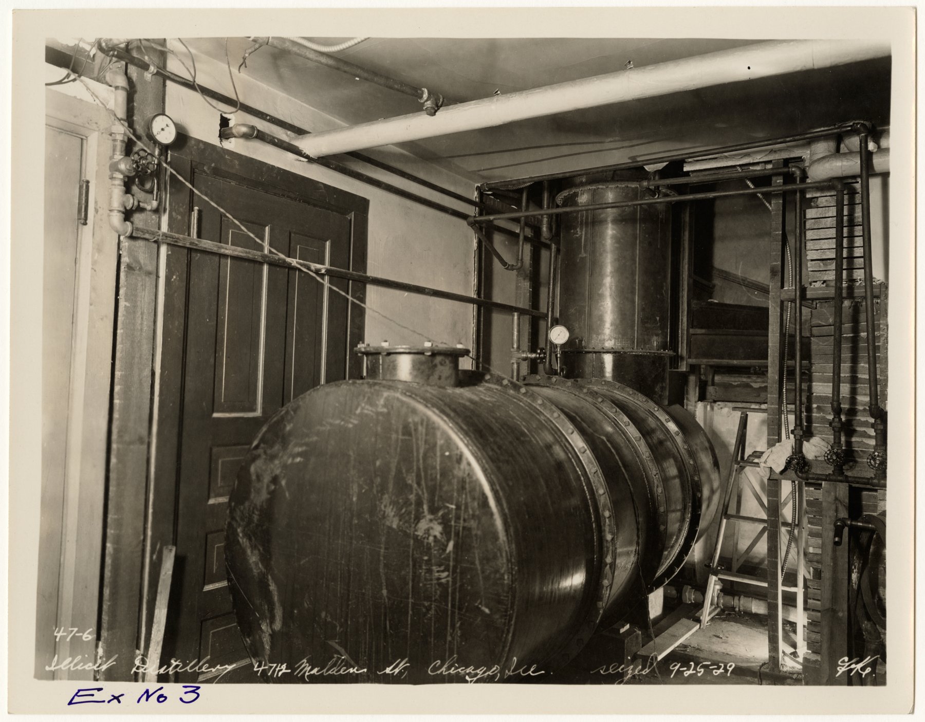 A large riveted horizontal tank and a vertical metal column with pipes, gauges and a ladder in a room beside a panelled door, with a handwritten caption reading Illicit Distillery, Chicago, Ill., seized 9-25-29, and Ex No 3 in blue.