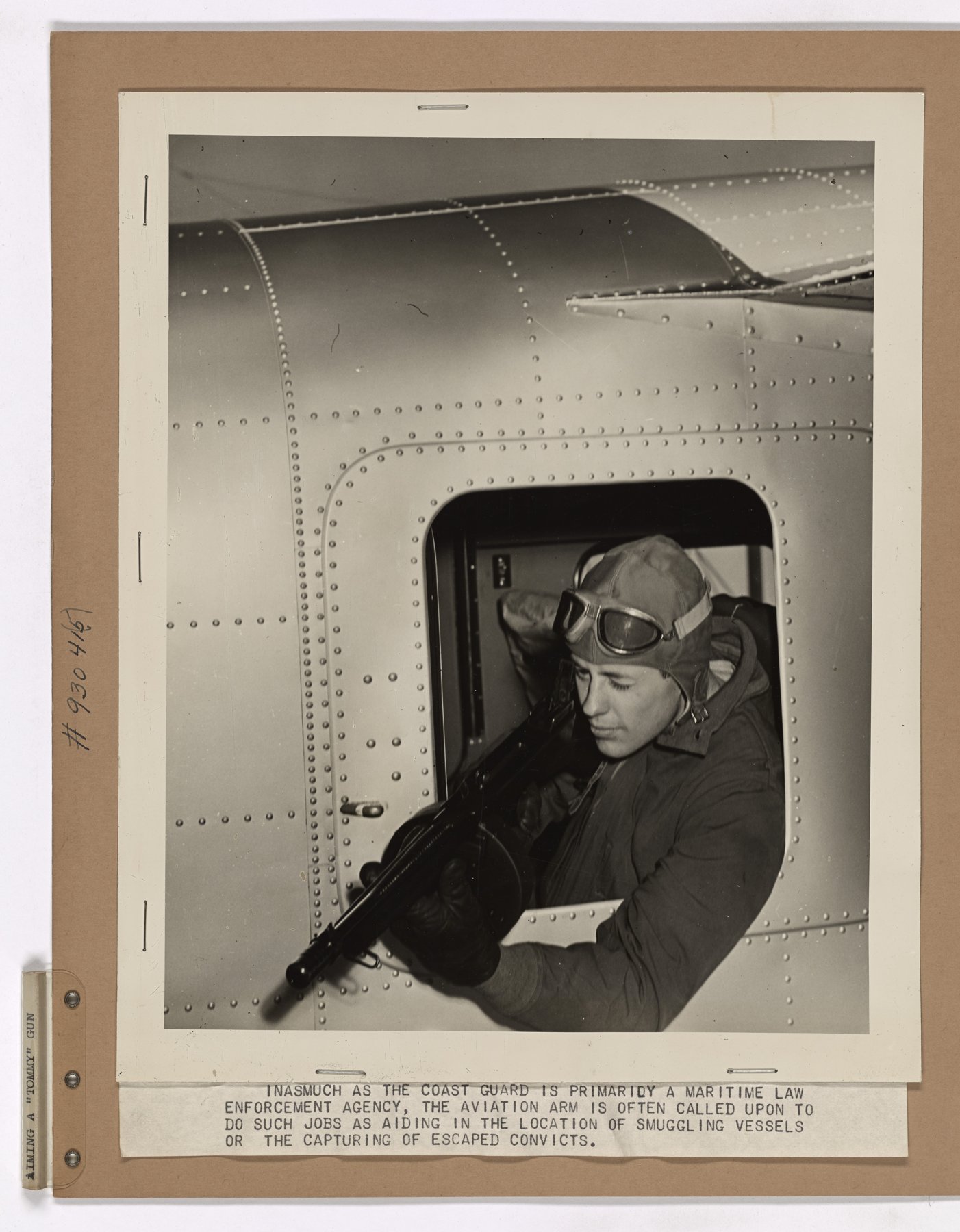 Mounted print of a man in a flying helmet and goggles aiming a submachine gun with a drum magazine out of a riveted aircraft window, with a typed caption below about the Coast Guard's aviation arm.