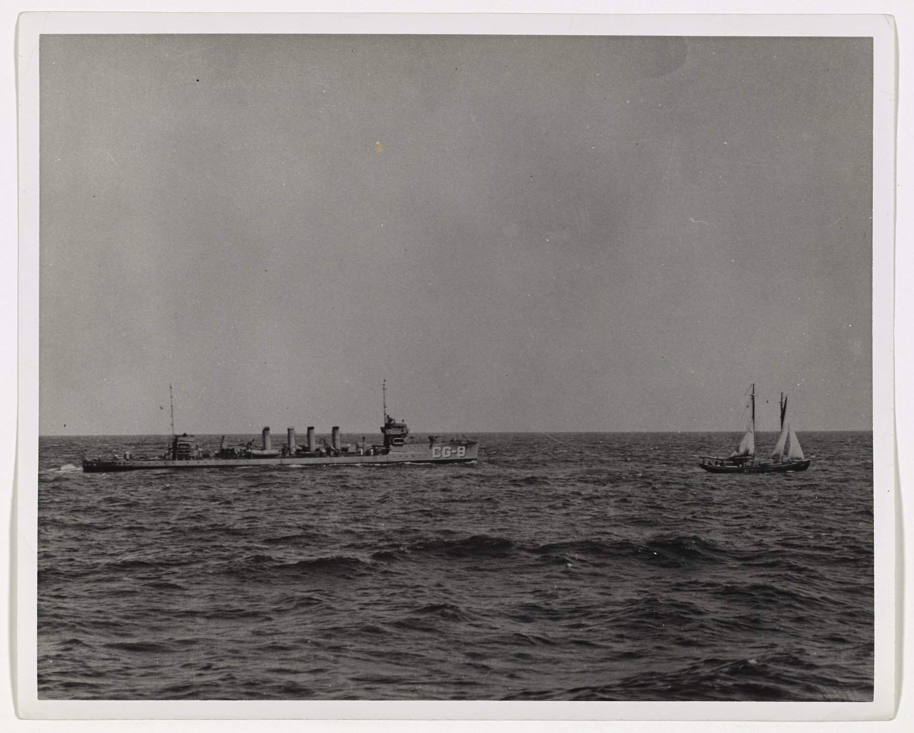 A four-stack destroyer far off on open sea at left, and a two-masted sailing vessel under sail at right.