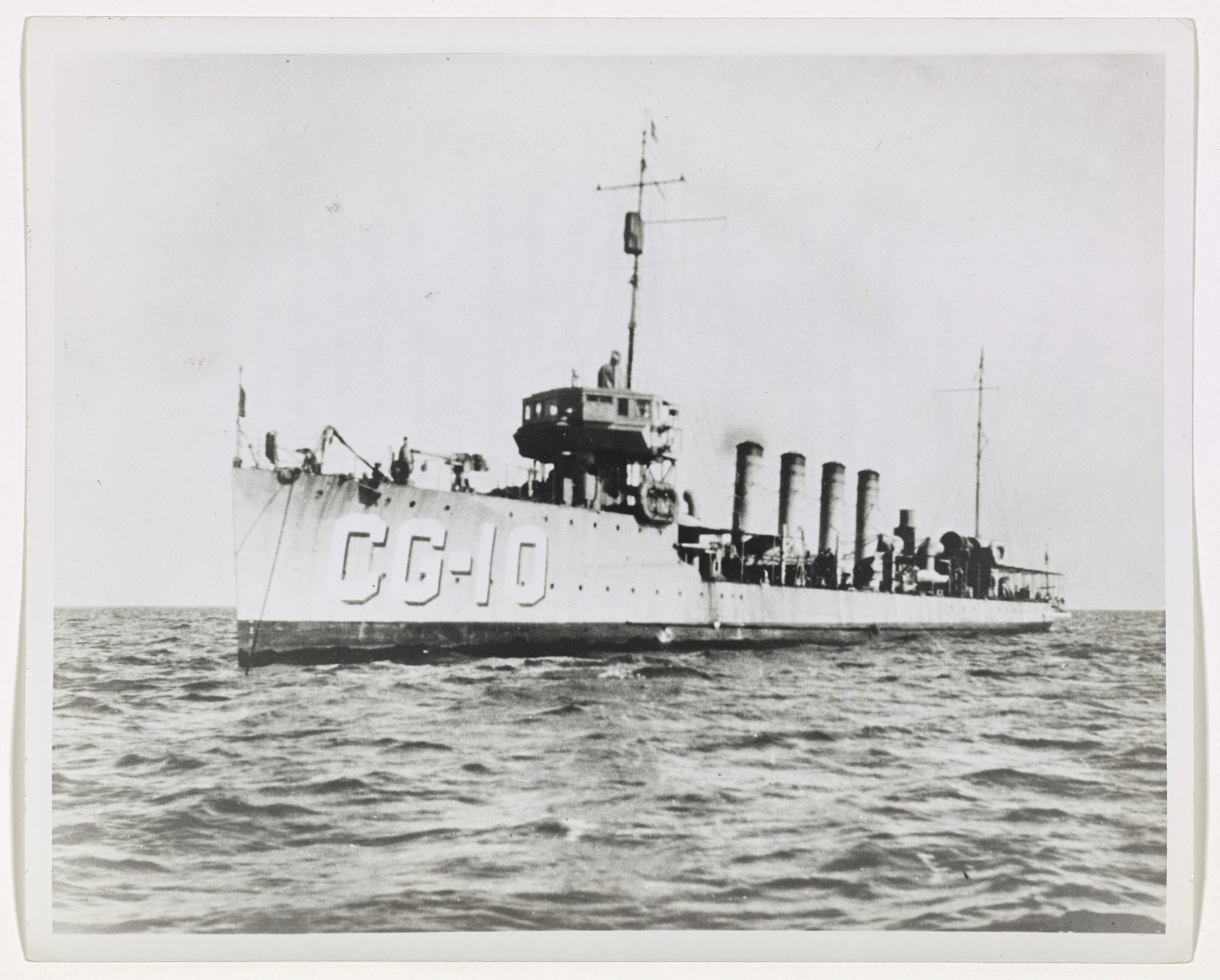 A four-stack destroyer seen bow-on at an angle, hull number CG-10, with a man on the bridge.