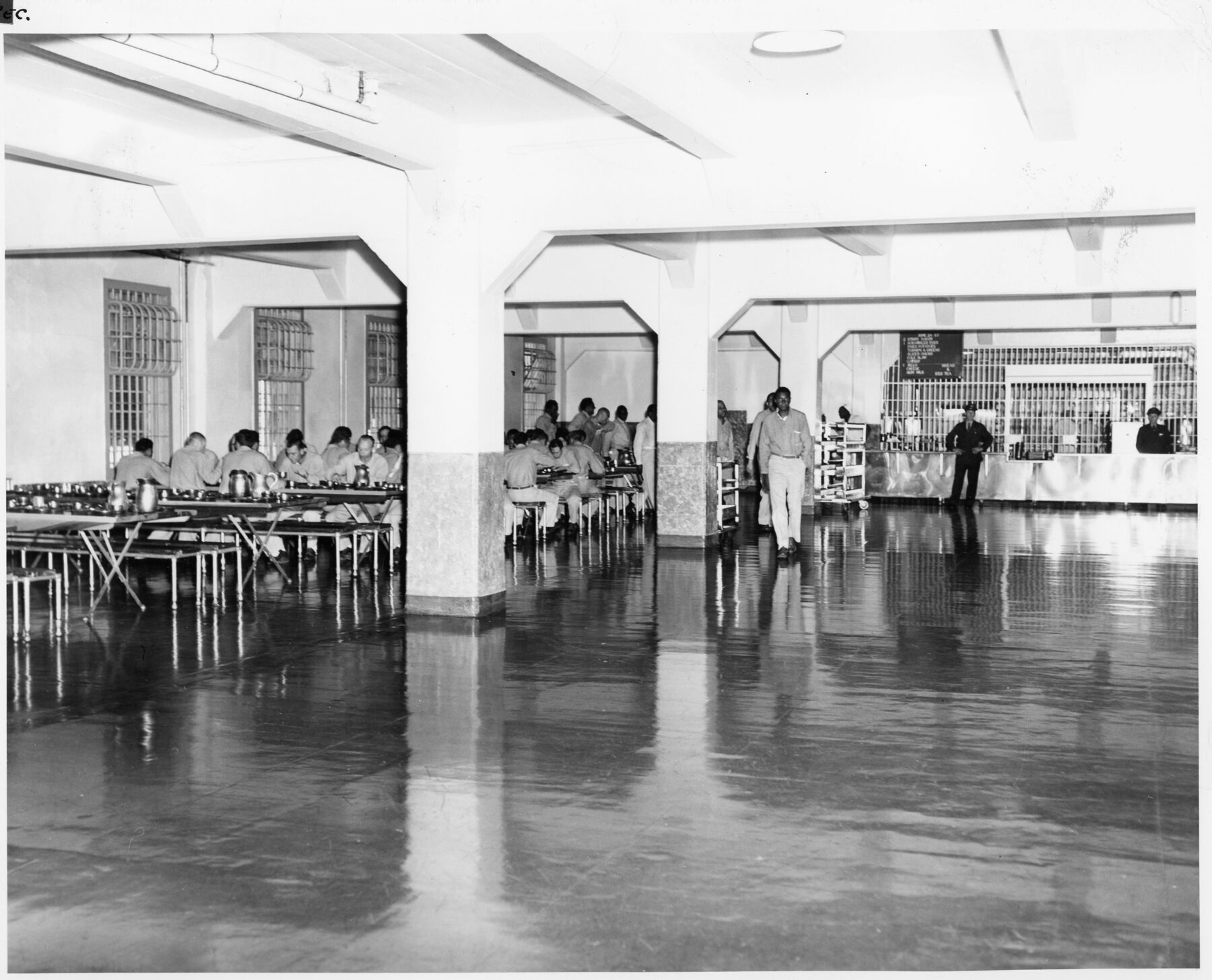 Black-and-white photograph of the Alcatraz dining room, with inmates seated at tables at the left under a low ceiling and columns, and staff standing near a serving area at the right.