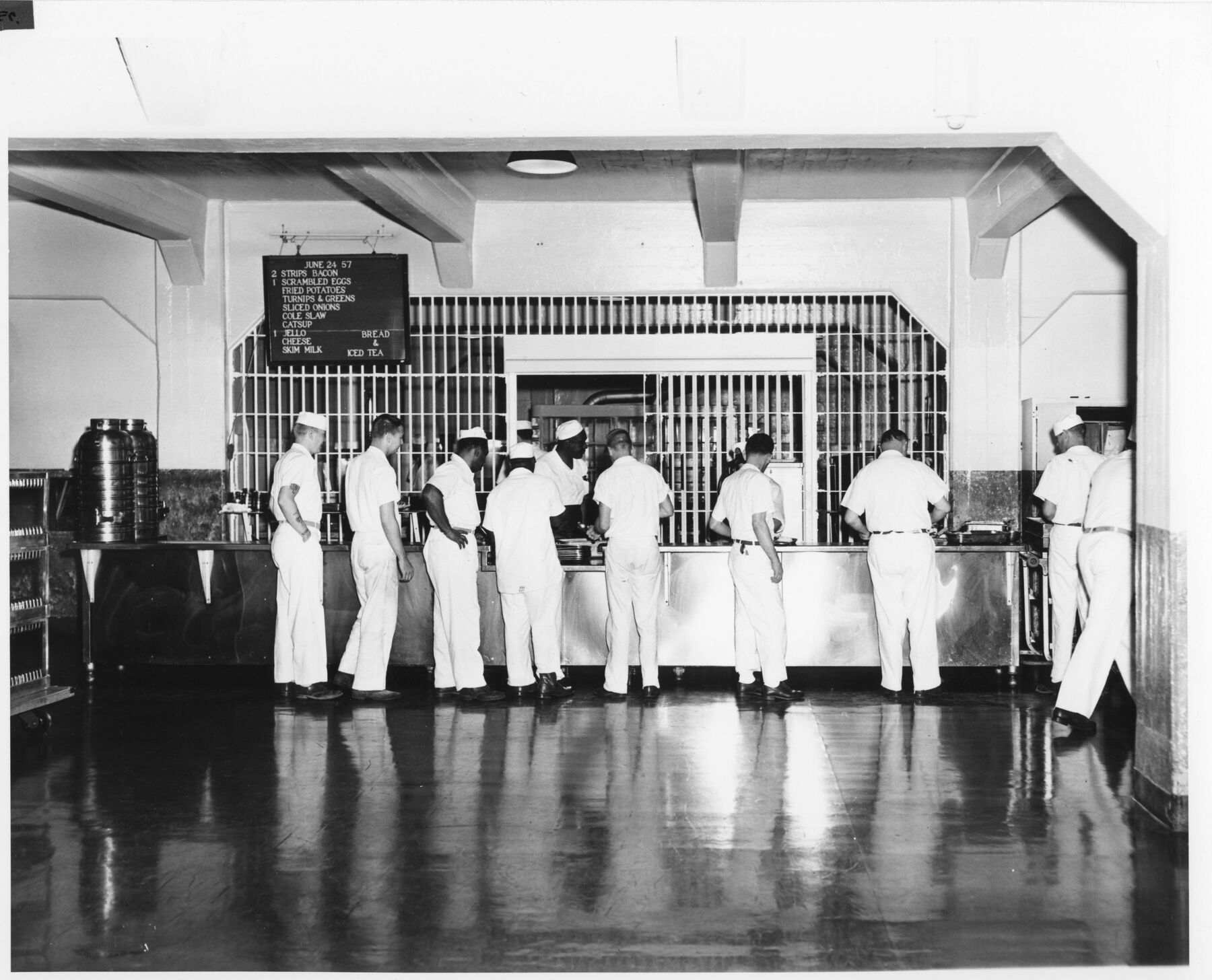 Black-and-white photograph of men in white uniforms and caps standing at a steel serving counter behind a barred screen, with a menu board above and a large urn at the left.