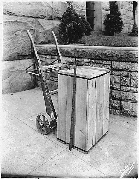A wooden crate bound with metal straps standing on pavement beside a hand truck, with a measuring stick leaning against it and a stone wall and shrubs behind.