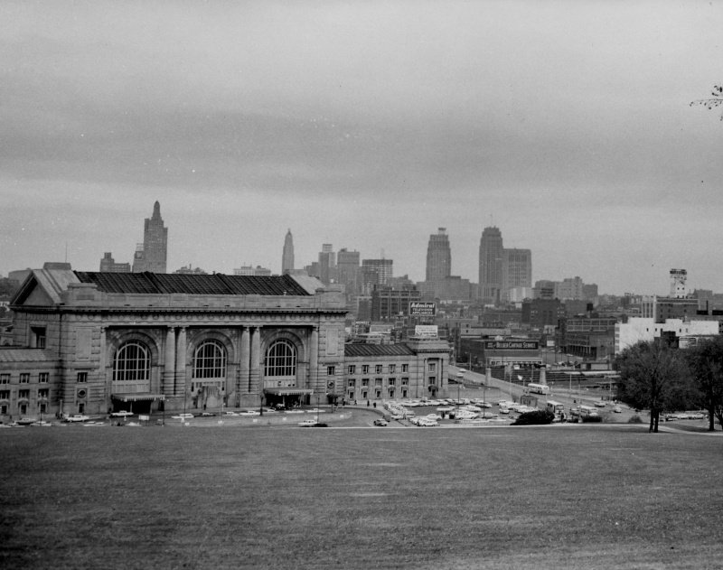 Wide view across a lawn of a large stone railway station with three tall arched windows, parking lots full of cars in front and the downtown Kansas City skyline behind.