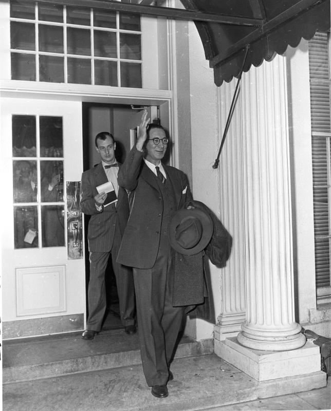 Senator Estes Kefauver in glasses, carrying a hat and coat and raising one hand, on the steps of the White House, with a man holding papers in the doorway behind him.