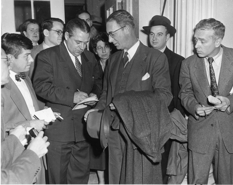 A tall man in glasses with a coat and hat over his arm, surrounded by reporters writing in notebooks.