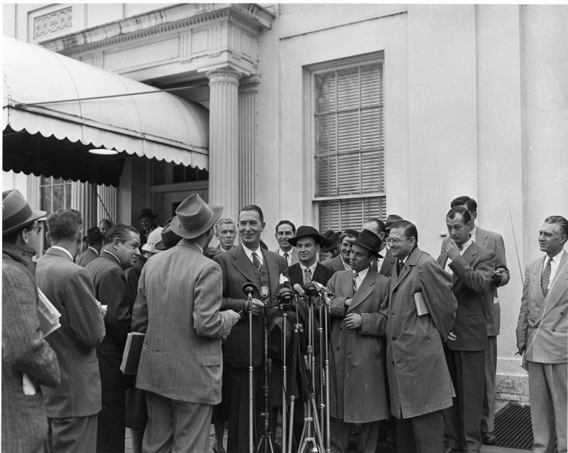 A group of men outside a building entrance with an awning and columns; a tall man in the centre smiles at a cluster of microphones as reporters with notebooks crowd around.