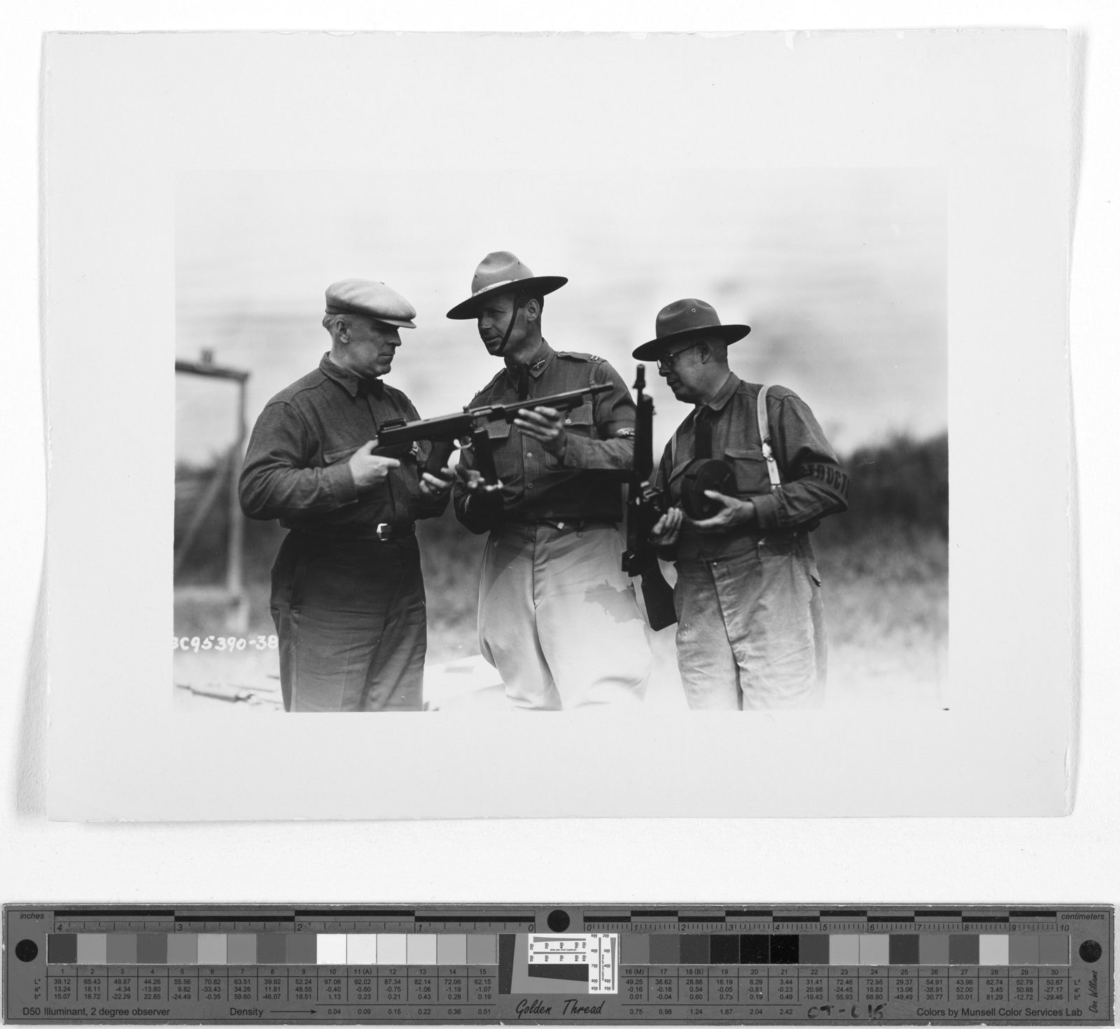 Three men outdoors at a rifle range. The man on the left, in a flat cap, and the Army officer in the center, in a campaign hat, hold a Thompson submachine gun between them; the man on the right holds a second Thompson upright and a drum magazine.