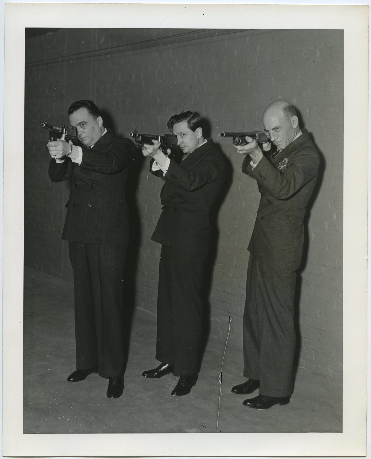 Three men in dark suits standing against a brick wall, each aiming a shoulder-fired gun to the left of the camera.