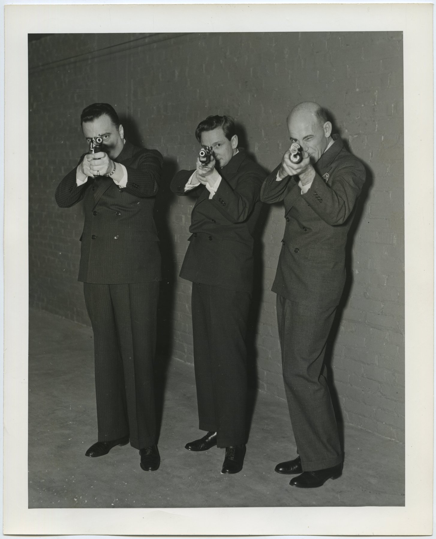 Three men in suits against a brick wall, each aiming a handgun toward the camera.