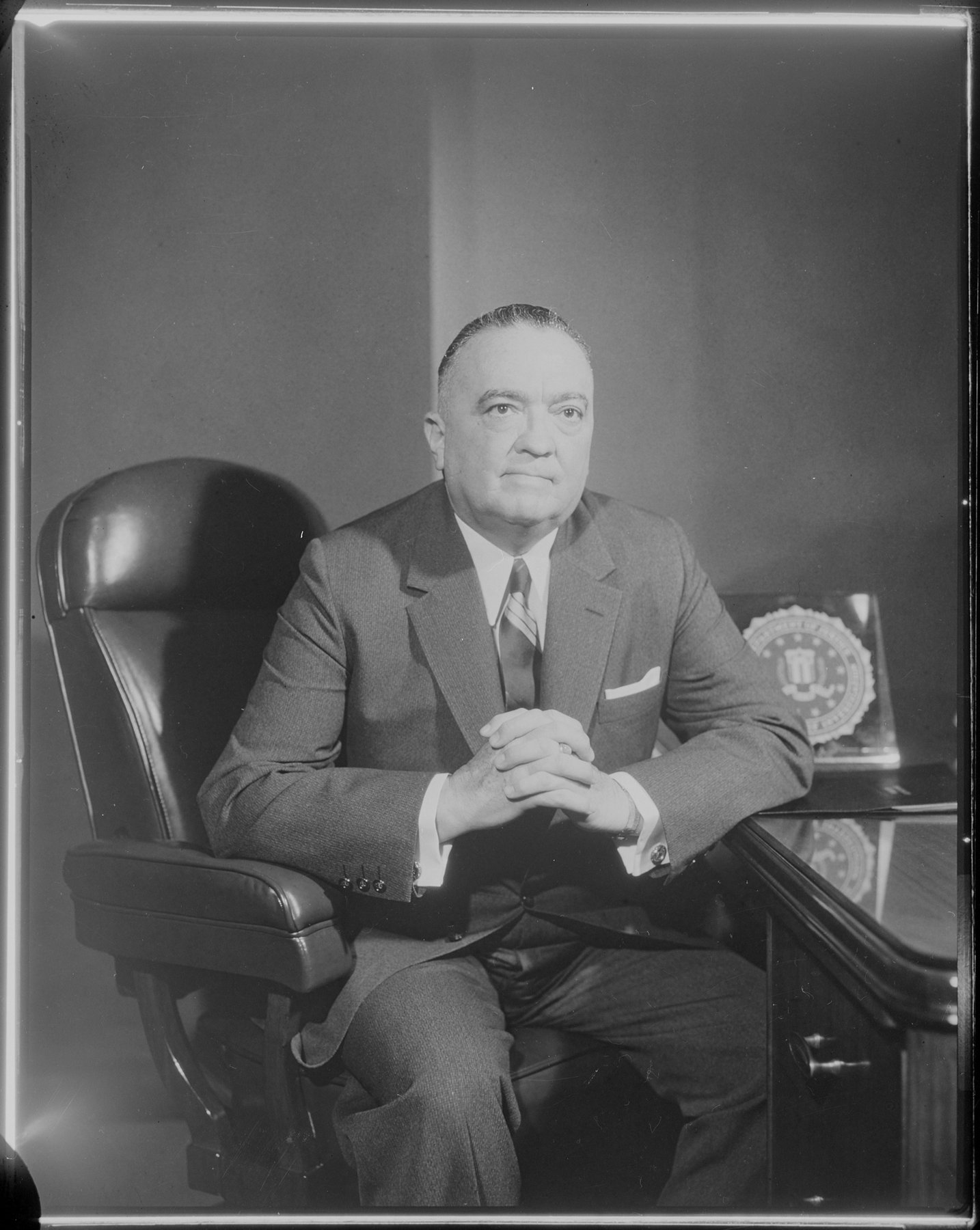 Studio portrait of a heavy-set man in a suit, striped tie and pocket square, seated in a leather chair with his hands clasped on a desk beside a plaque bearing the FBI seal.