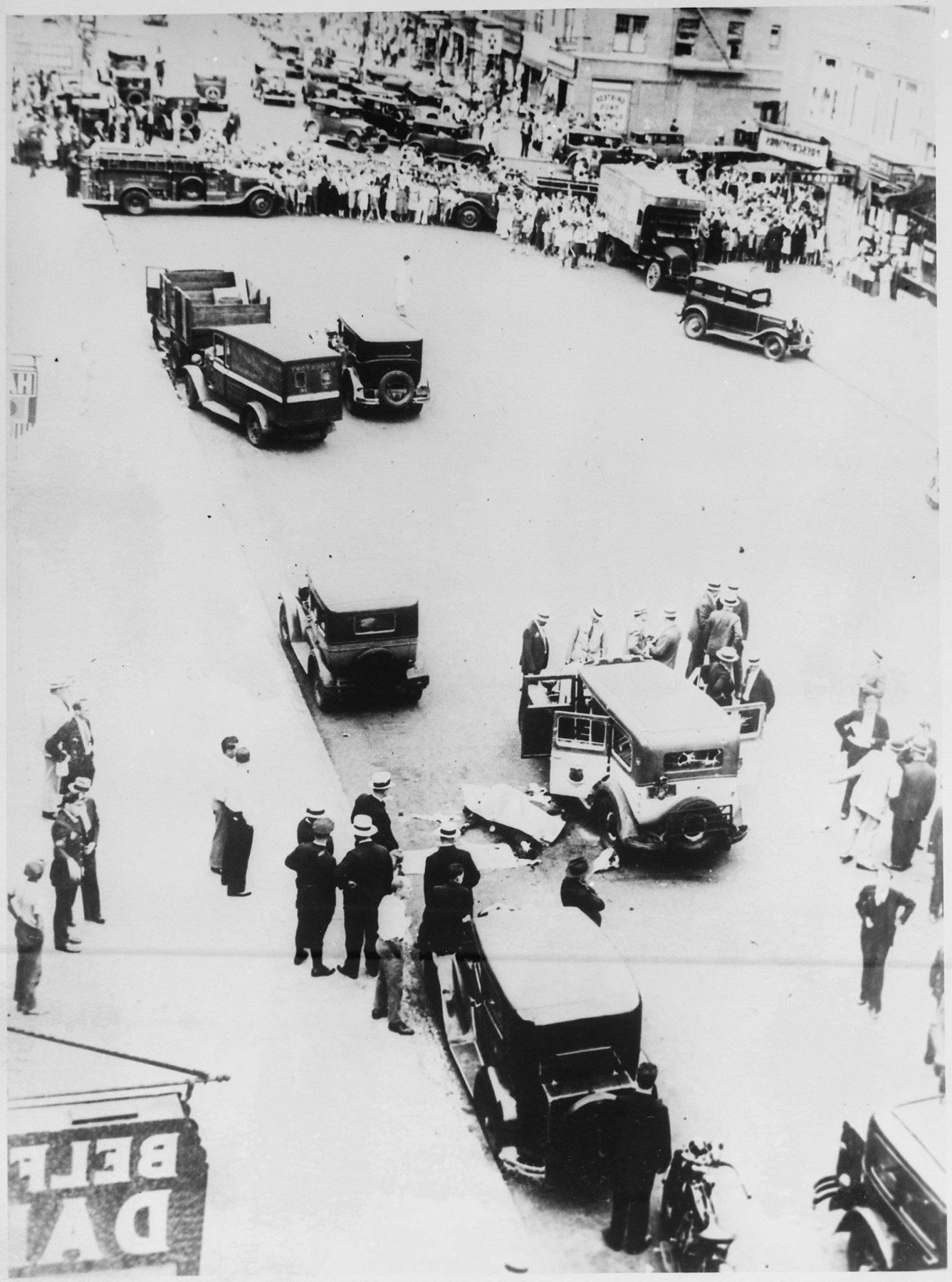 High view of a city street: a taxi with its doors open, a covered form on the pavement beside it, men in straw hats and uniformed police around it, and a fire engine and crowd held back at the far end.