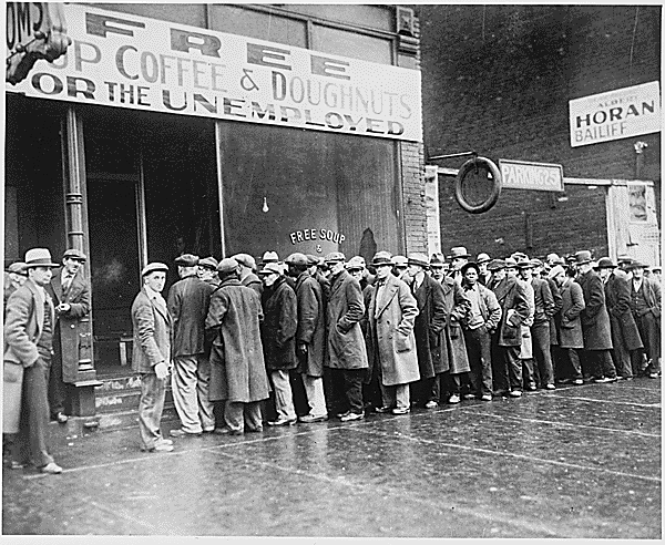 A line of men in overcoats and hats along a city sidewalk outside a storefront with a large sign reading Free Soup, Coffee and Doughnuts for the Unemployed.
