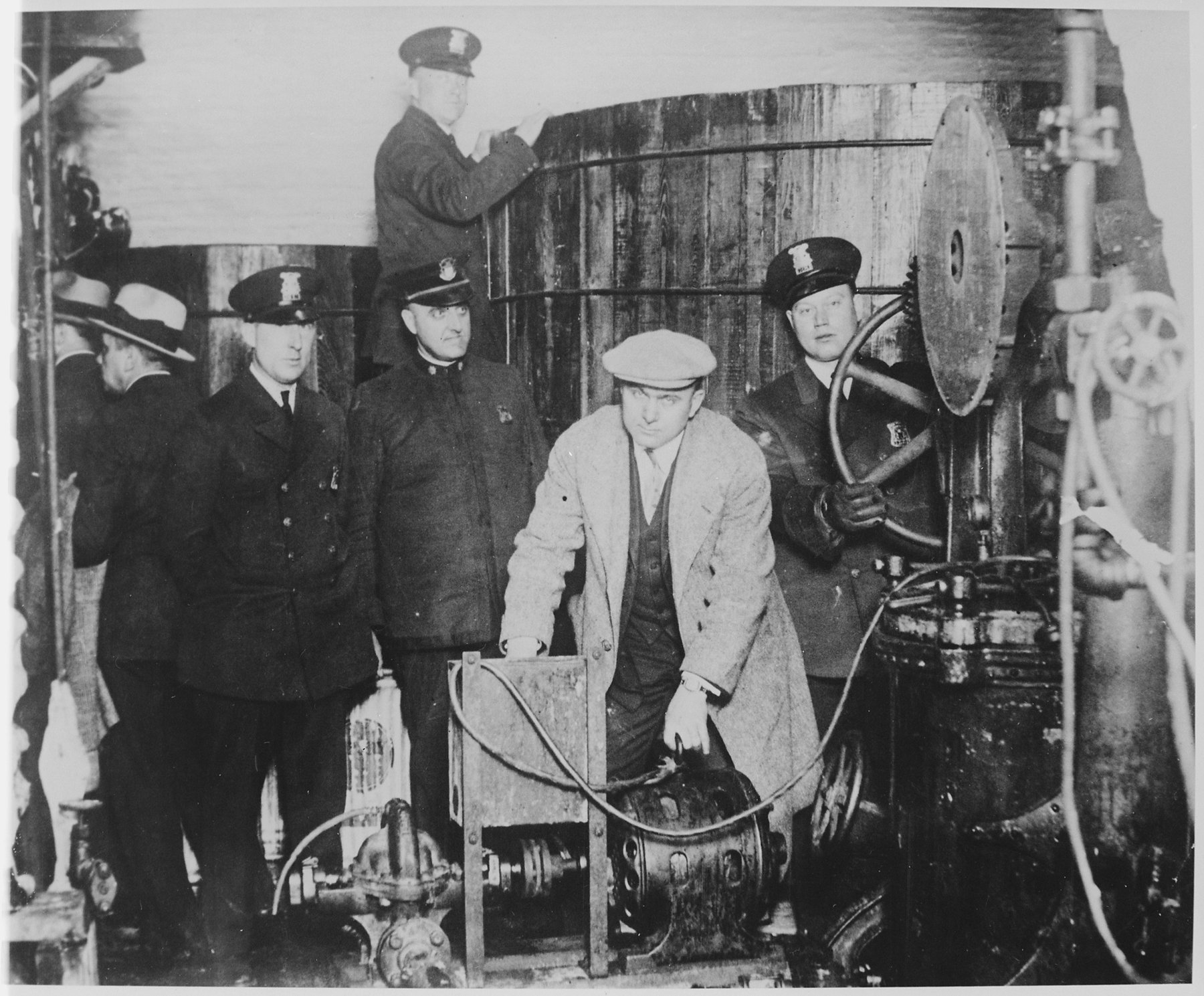 Uniformed police in peaked caps and men in caps and hats beside a large wooden vat, a motor-driven pump with a valve wheel and pipes; one man in a flat cap crouches at the motor.