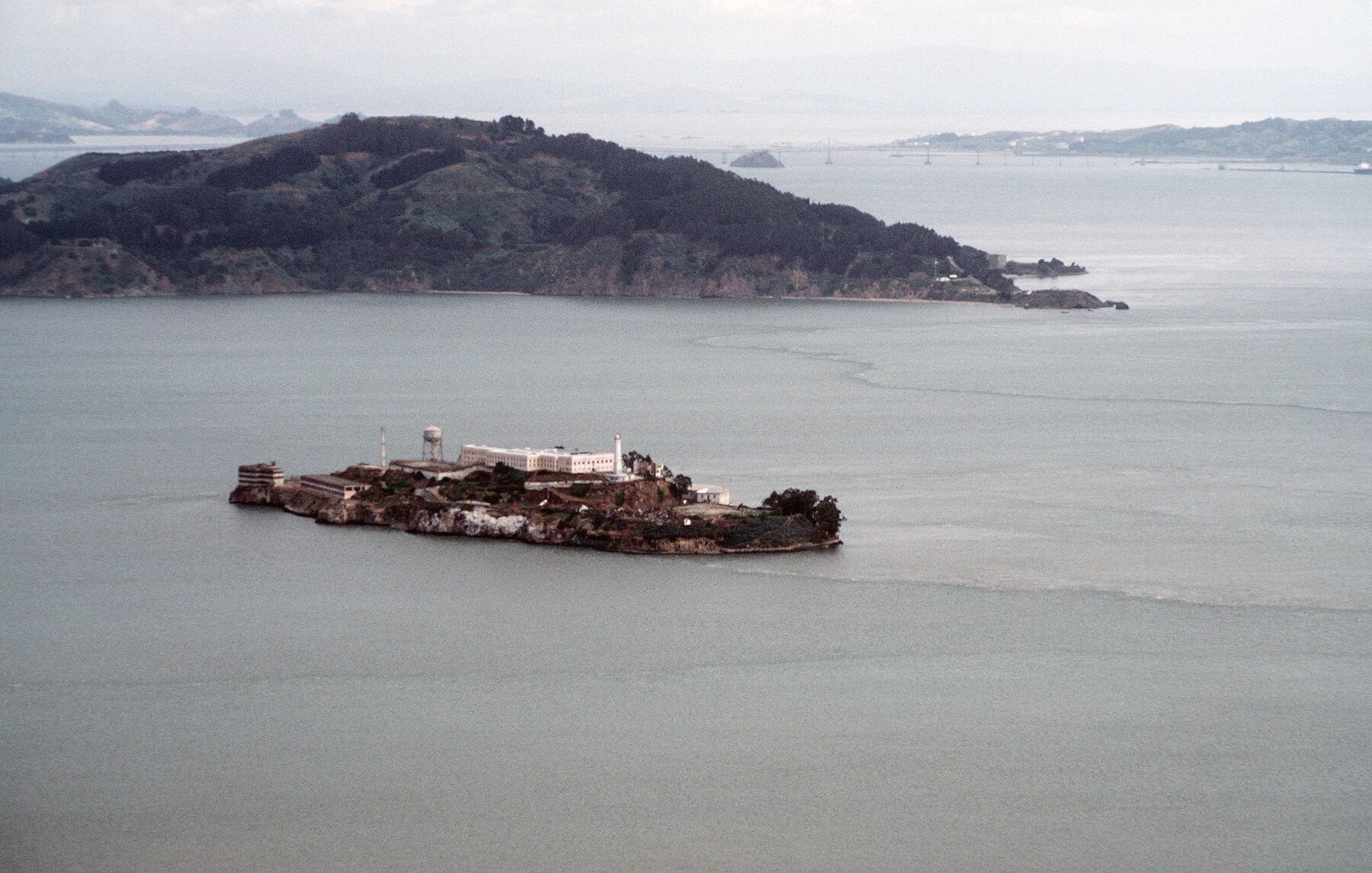 Color aerial photograph of Alcatraz Island in San Francisco Bay, with Angel Island and the Marin shoreline behind it.