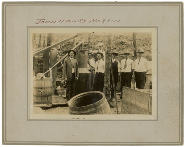 Mounted photograph headed in handwriting John Henry Hardin, showing seven men in hats standing by a still with a tall metal column, a pipe and barrels, one barrel overturned in front, in woods.