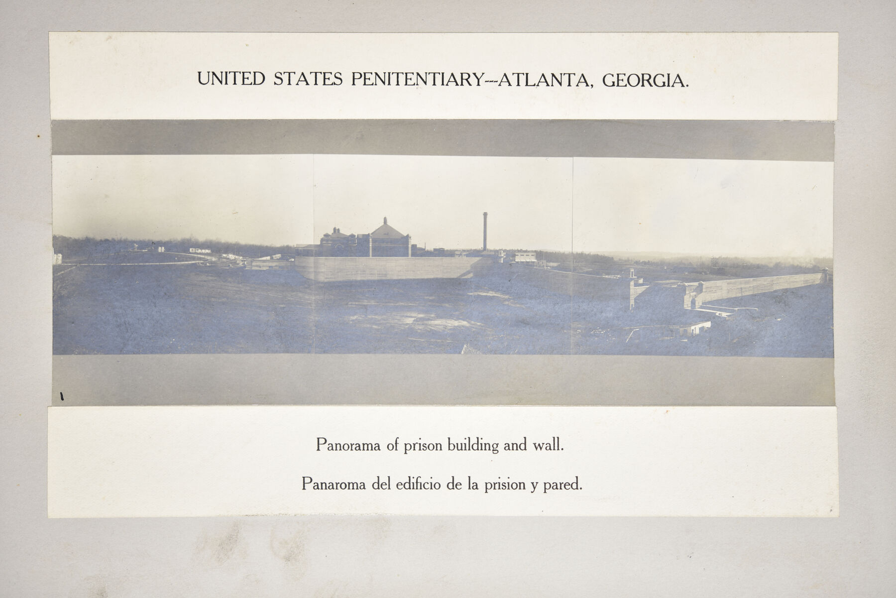 Mounted panorama strip of open ground with a distant prison building, a chimney and a wall at the right, above an English and Spanish caption.