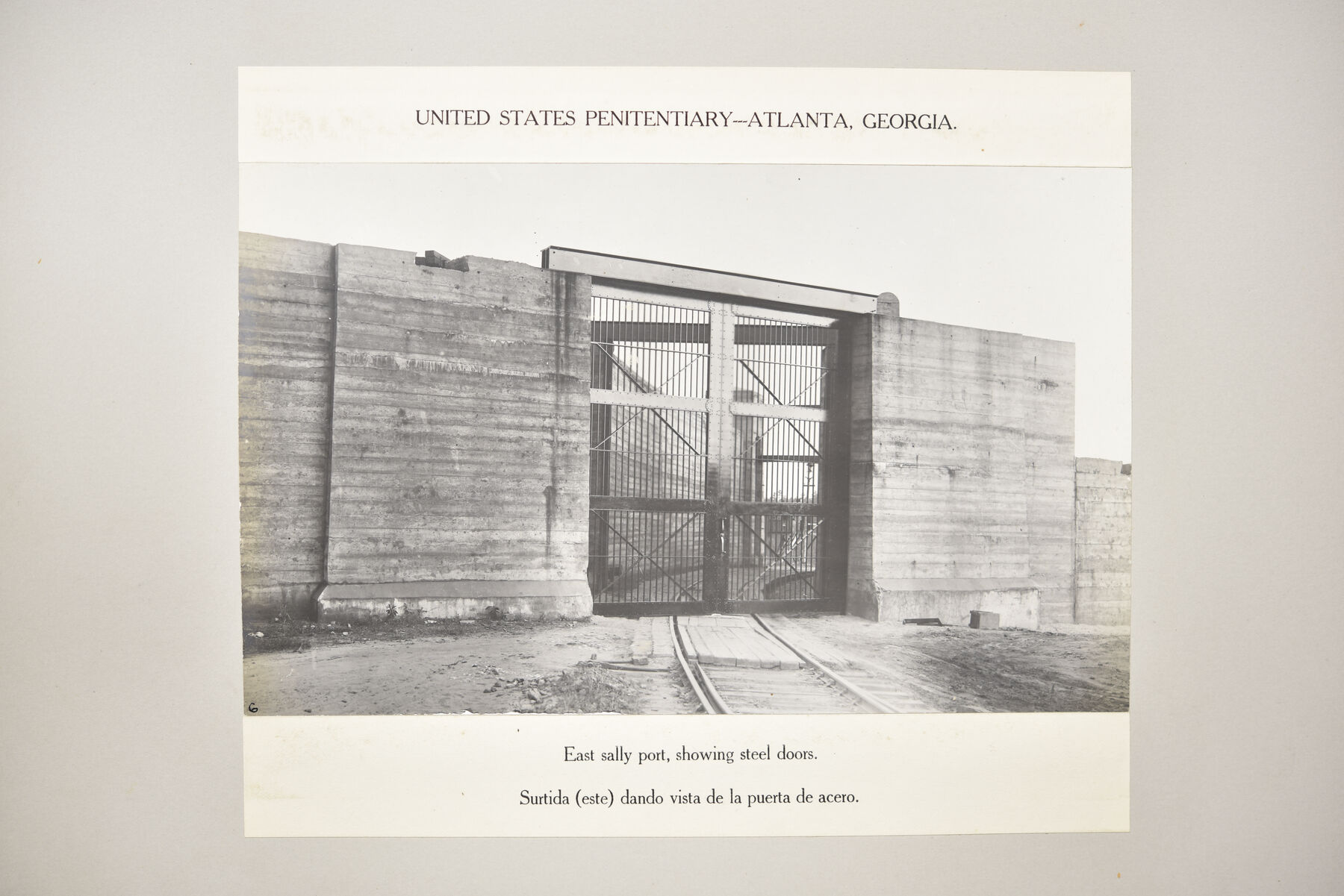Mounted photograph of a concrete wall pierced by a barred steel gate with a rail track in the foreground.