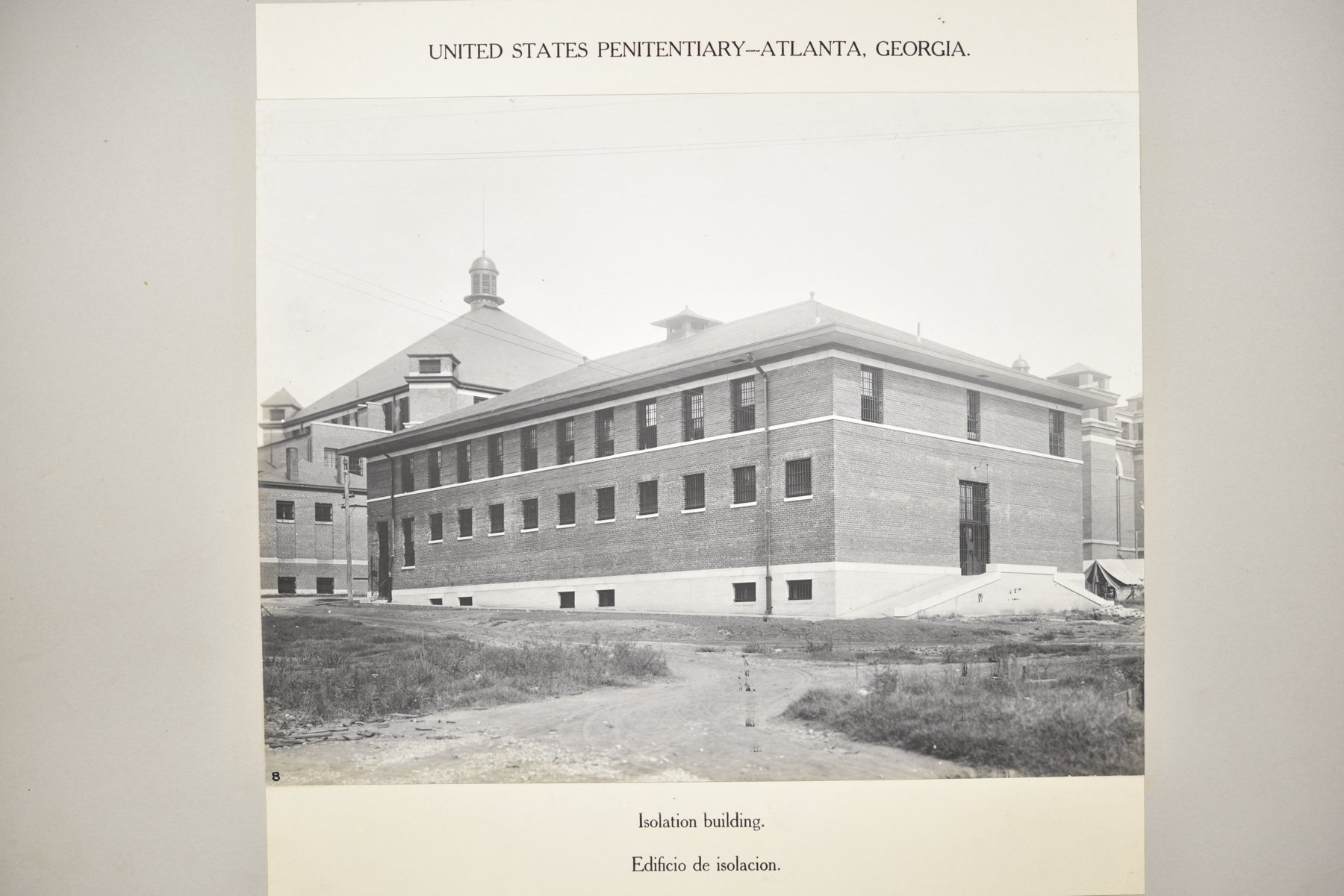 Album page headed United States Penitentiary, Atlanta, Georgia, with a photograph of a two-storey brick building with barred windows on bare ground, with the domed cell house behind it, above the caption Isolation building.