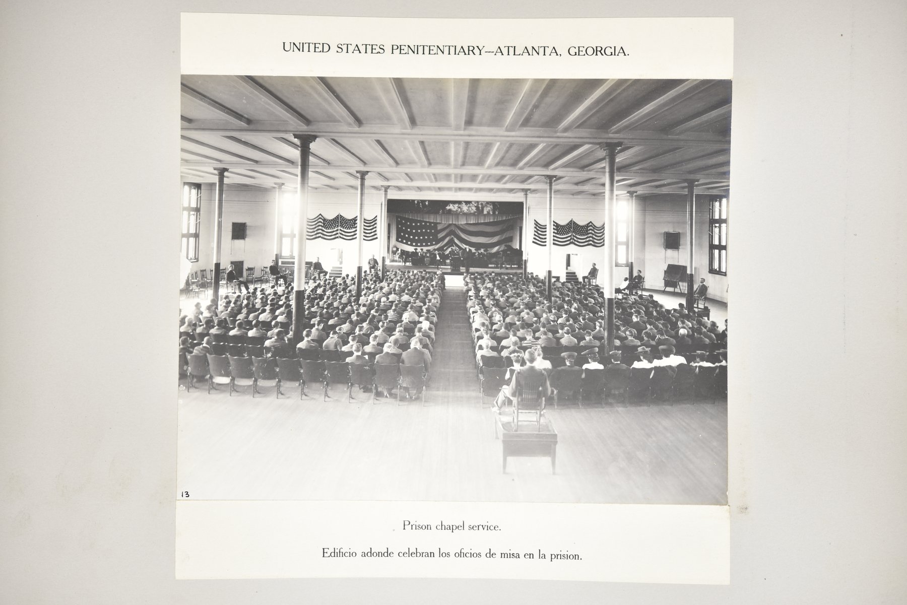 Album page headed United States Penitentiary, Atlanta, Georgia, with a photograph of a large hall with columns, flags draped across the far wall above a stage, and rows of seated men facing the stage, with a few men seated at the sides and a chair on a low platform in the aisle, above the caption Prison chapel service.