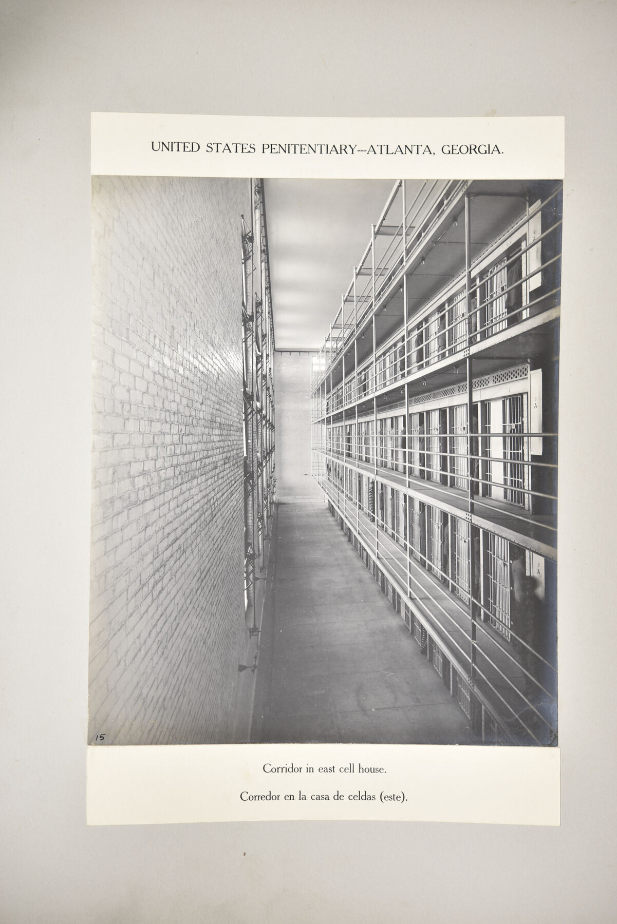 Mounted portrait-format photograph looking down a long corridor of the east cell house at Atlanta, with a brick wall at the left and tiers of cells at the right.