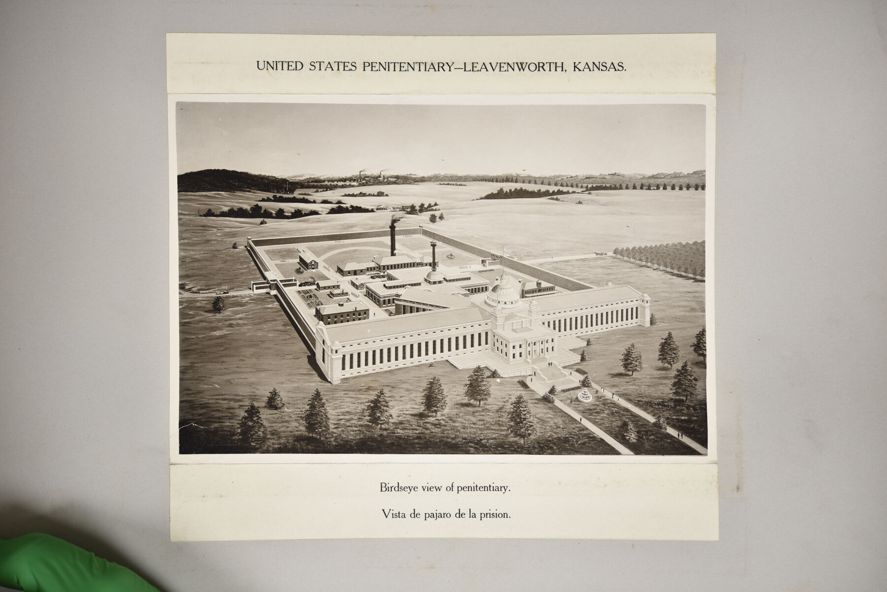 Mounted print showing an illustrated bird's-eye view of the Leavenworth penitentiary complex under a typed caption, with a green glove at the lower left edge of the scan.