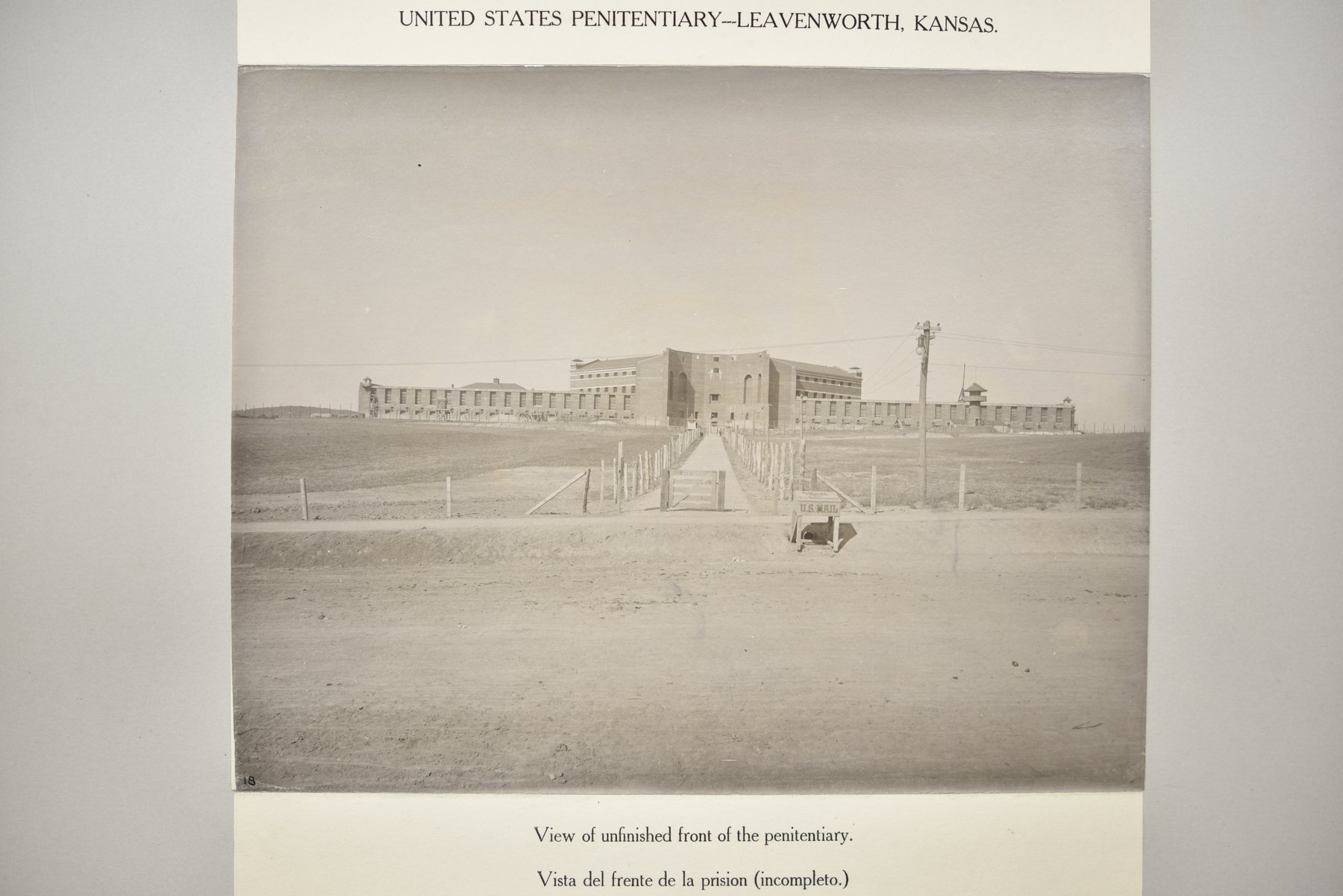 Album page headed United States Penitentiary, Leavenworth, Kansas, with a photograph of The photograph looks down a fenced walk across open ground to the long front of the penitentiary, with a taller central block, a guard tower at the right, a telegraph pole and a small sign by a dirt road in the foreground, above the caption View of unfinished front of the penitentiary.