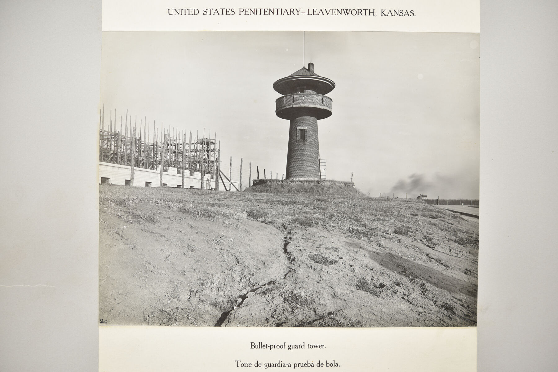 Mounted photograph of a dark round guard tower with a roofed lookout on bare ground, with construction scaffolding of a wall behind it.
