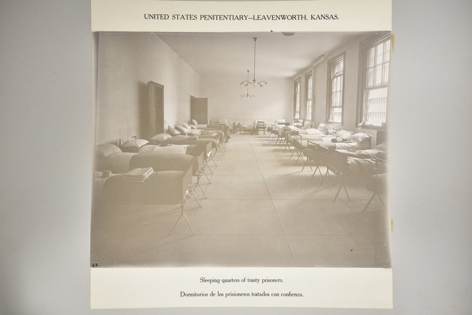 Album page headed United States Penitentiary, Leavenworth, Kansas, with a photograph of a long, high room with tall windows on the right, rows of made iron cots along both walls, and hanging lamps; no one is in the room, above the caption Sleeping quarters of trusty prisoners.