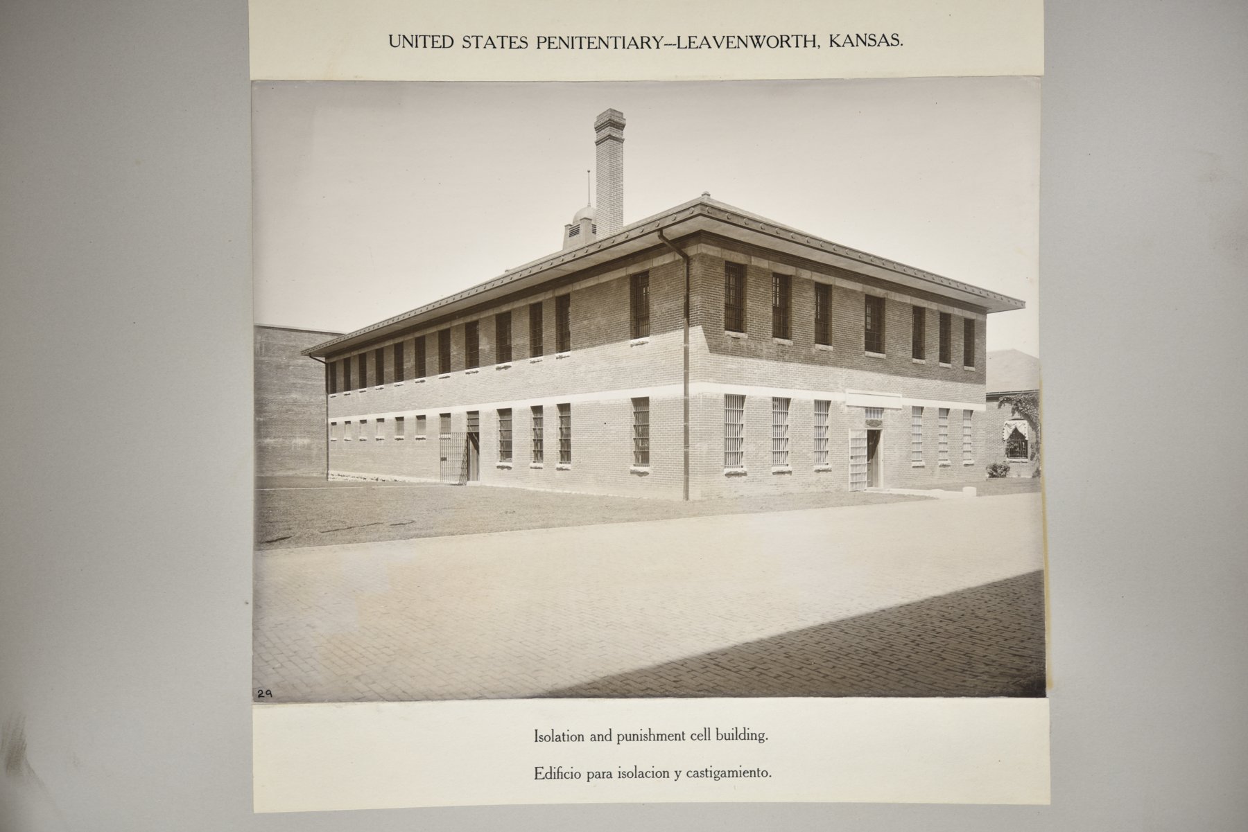 Album page headed United States Penitentiary, Leavenworth, Kansas, with a photograph of a two-storey brick building with barred windows, a hipped roof and a chimney, over brick paving, above the caption Isolation and punishment cell building.