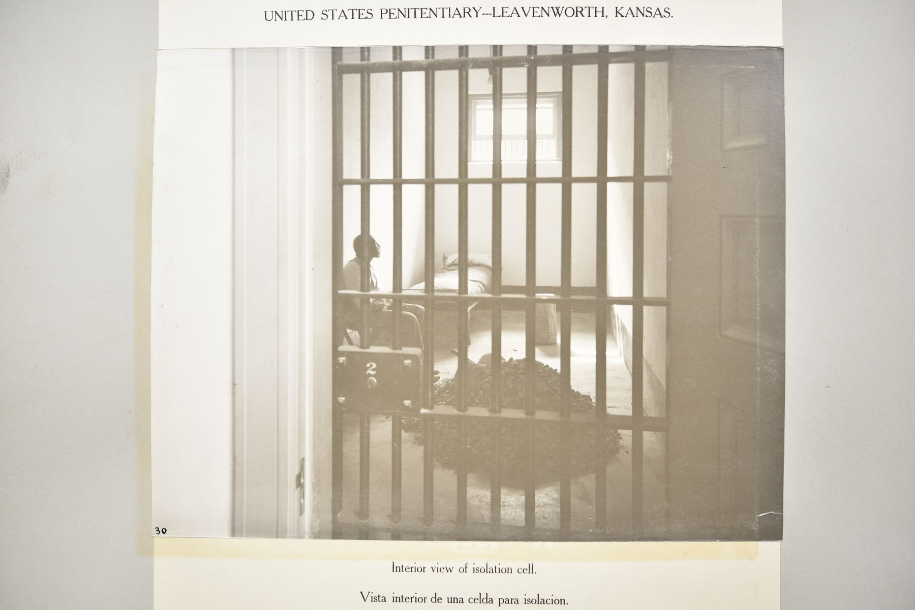 Mounted photograph taken through cell bars into a small isolation cell with a bunk and a figure inside.
