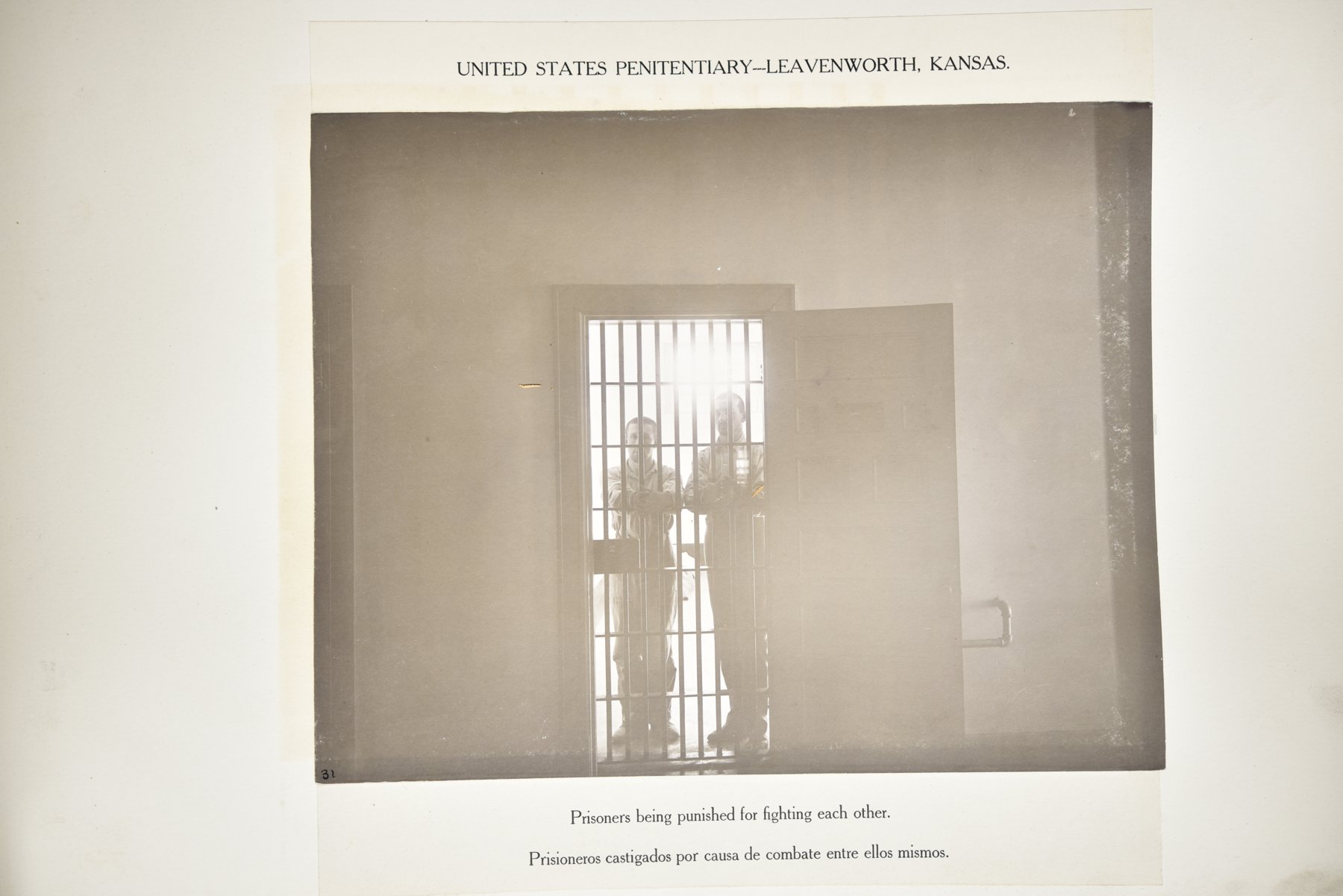 Album page headed United States Penitentiary, Leavenworth, Kansas, with a photograph of two men in light uniforms standing behind a barred cell door, backlit, with the door open outward, above the caption Prisoners being punished for fighting each other.